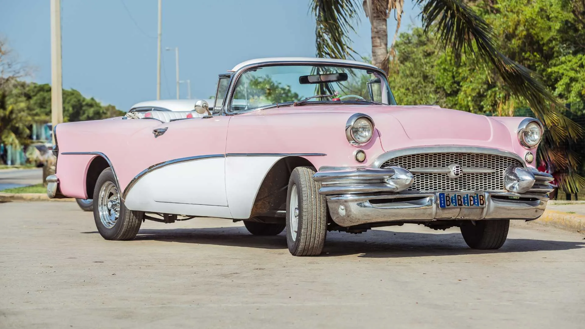 A vintage pink and white convertible car parked on a sunny street near palm trees.