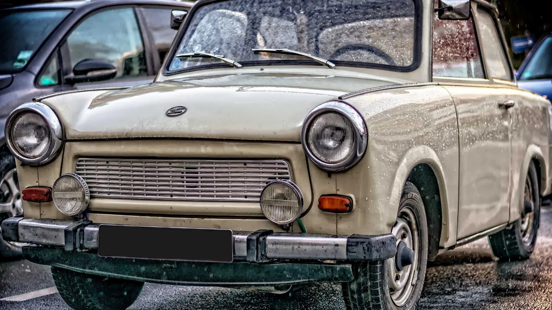 A vintage yellow Trabant car parked in the rain, showcasing its classic design with a chrome bumper and round headlights.