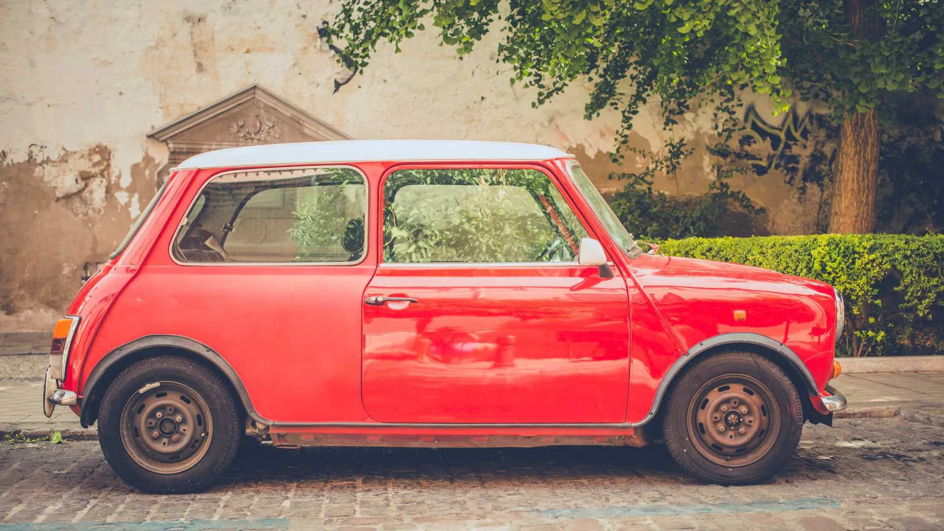 Red vintage Mini Cooper parked on a cobblestone street against an aged wall with greenery in the background.