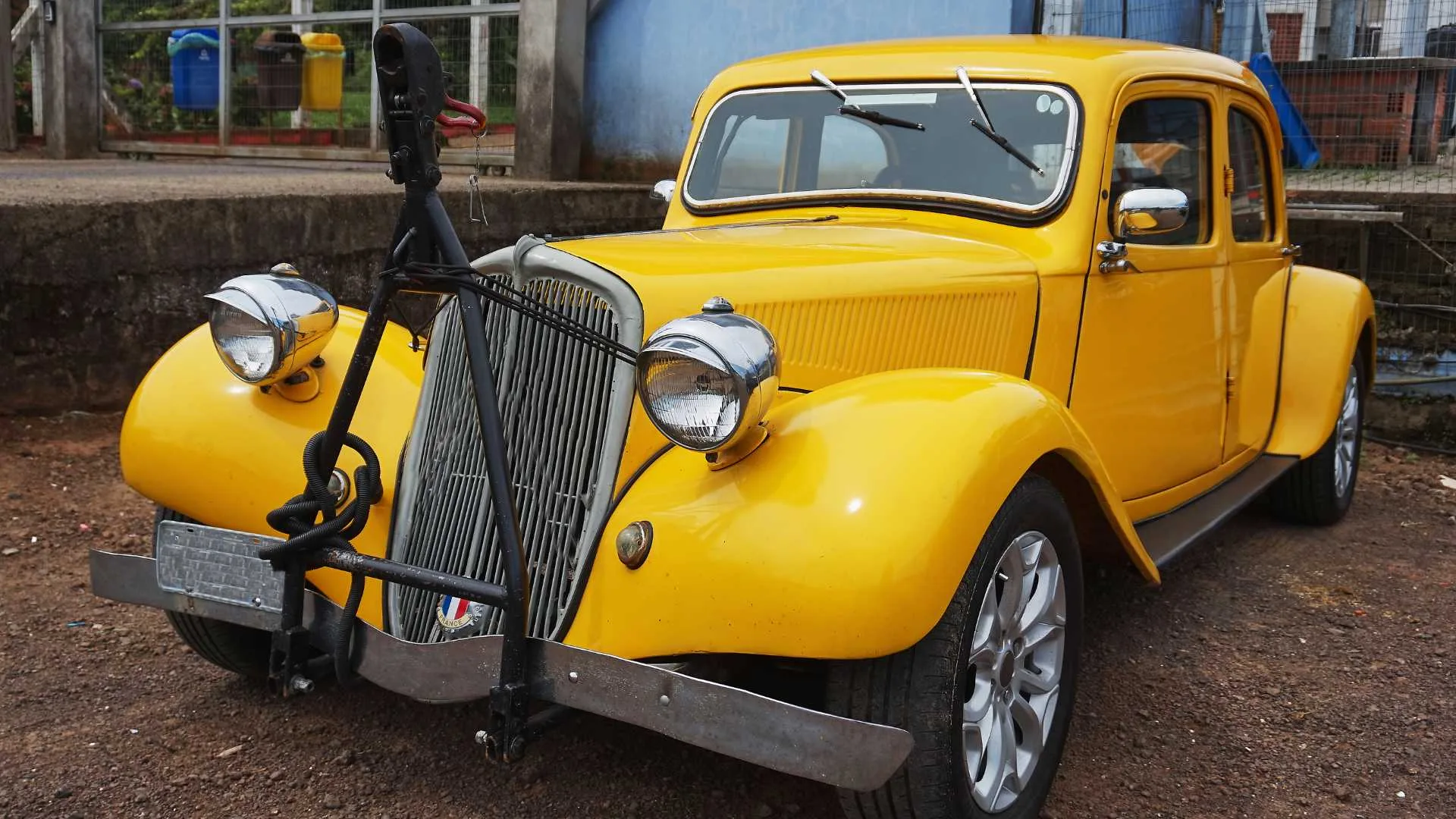 Yellow vintage car with a chrome grille and modern wheels parked on a dirt surface.